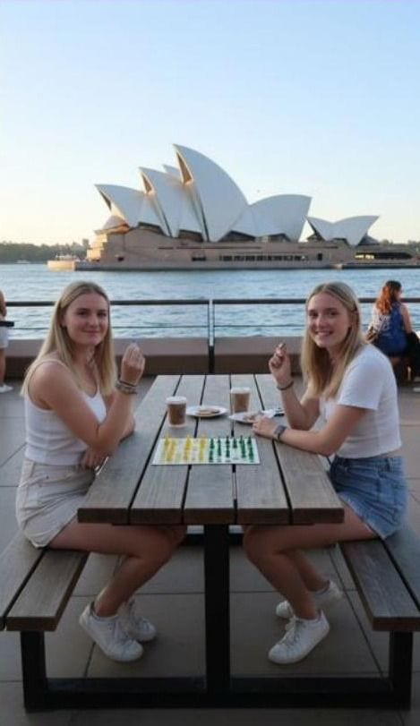 Two friends playing Aussie Checkers at an outdoor table beside Sydney Harbour, with the Opera House behind them.
