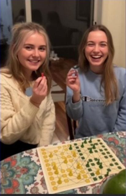 Two friends laughing over an Aussie Checkers board at home, pieces in hand.